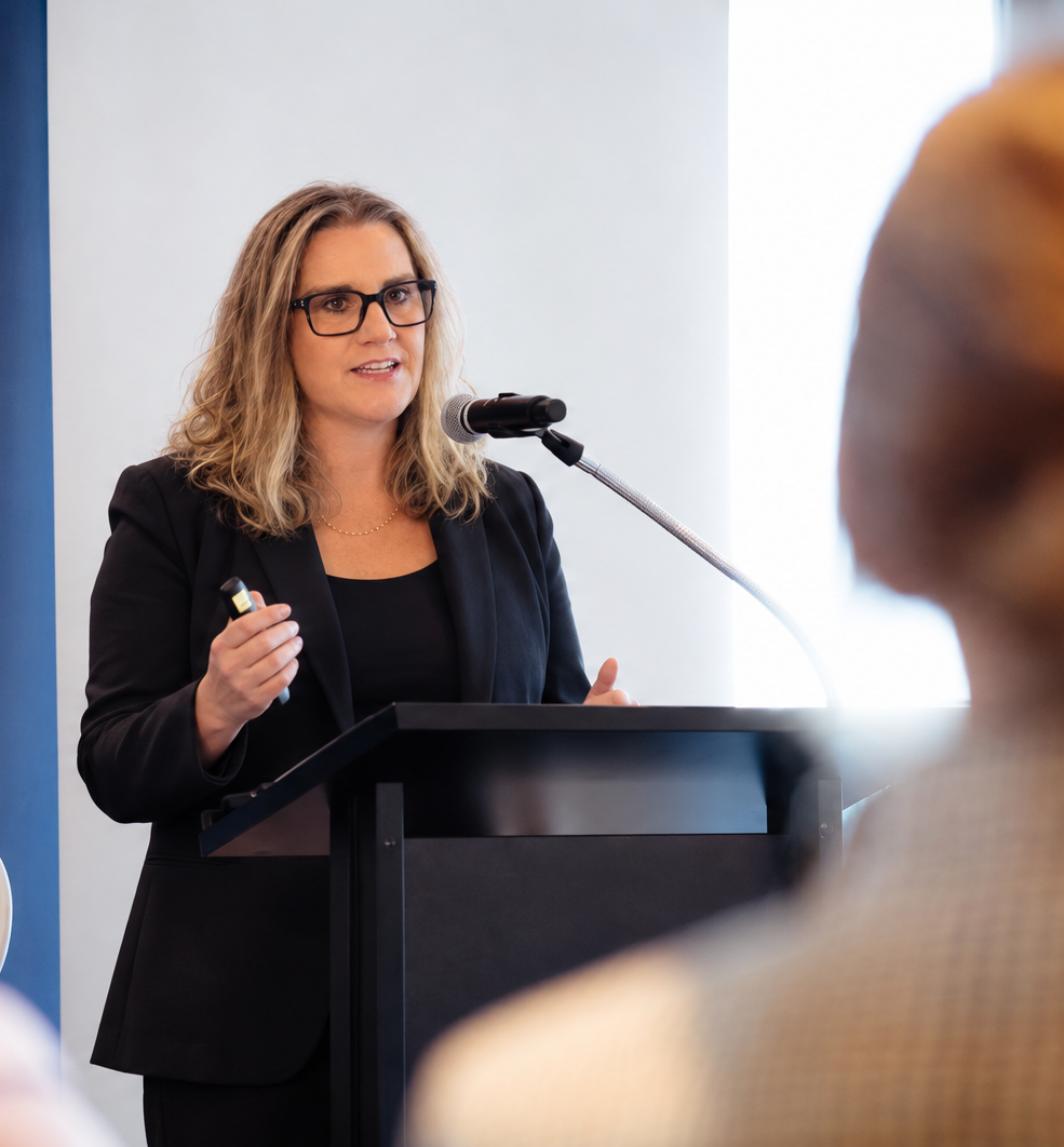 Claire Allison speaking at a lectern with a microphone, an audience member in the foreground